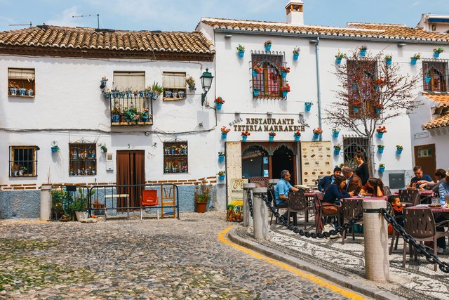 Marokkanischen Restaurant 'Tetería Marrakech' im Albaicín-Viertel von Granada, mit weiß getünchter Fassade, bunten Blumentöpfen an den Wänden und Gästen, die bei Sonnenschein auf dem Kopfsteinpflasterplatz traditionelle Speisen und Tee genießen.