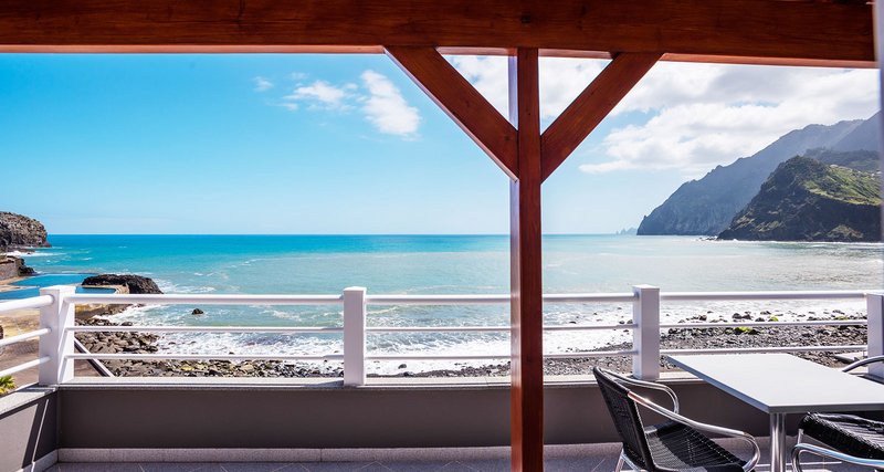 Terrassenblick auf den Atlantik in Porto da Cruz – wilde Küstenlinie, schwarzer Lavastrand und der markante Penha d’Águia im Hintergrund.