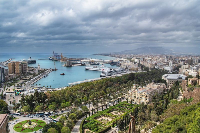Blick vom Castillo de Gibralfaro auf Málaga an einem bewölktem Tag, mit dem historischen Rathaus und seinen symmetrischen Gartenanlagen im Vordergrund sowie Kathedrale und Hafen im Hintergrund.