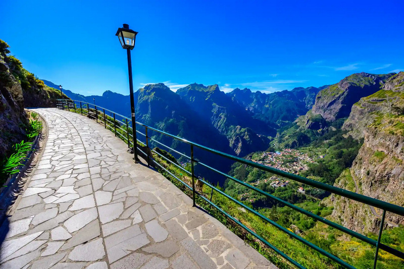 Gepflasterter Weg mit Laternen am Aussichtspunkt Eira do Serrado auf Madeira – Panoramablick ins grüne Curral das Freiras (Nonnental) mit steilen Bergflanken ringsum.