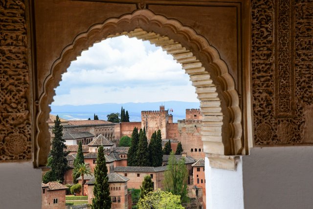 Blick durch ein reich verziertes maurisches Hufeisenfenster der Alhambra auf die Alcazaba mit Festungsmauern, Türmen und Zypressen vor der Bergkulisse Andalusiens