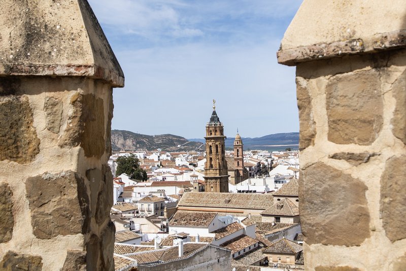 Blick durch die Zinnen der Alcazaba von Antequera auf die Altstadt mit der barocken Turmspitze der Kirche Real Colegiata de San Sebastián und umliegenden weißen Häusern.