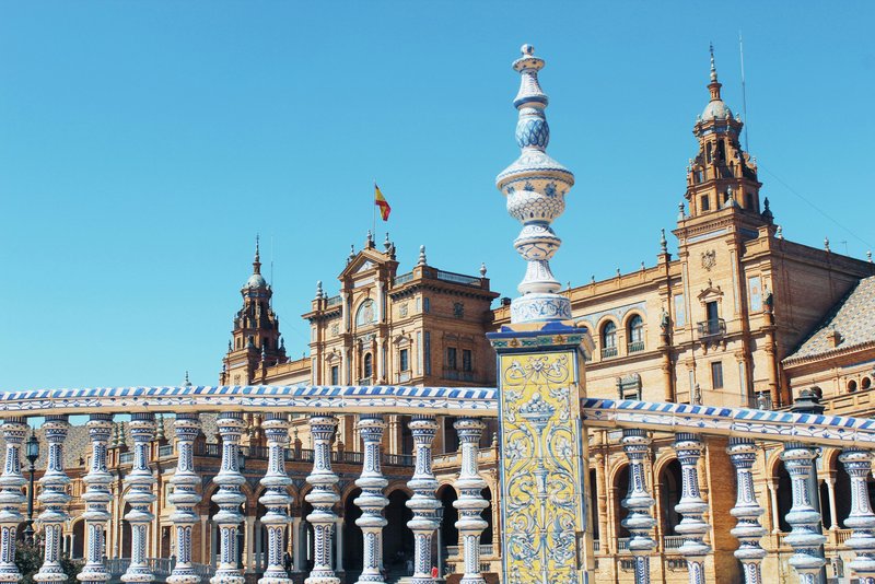 Nahaufnahme eines bunt glasierten Keramikgeländers auf einer Brücke am Plaza de España in Sevilla, mit Blick auf das Hauptgebäude im Regionalstil und der spanischen Flagge unter blauem Himmel – ein Wahrzeichen der Ibero-Amerikanischen Ausstellung von 1929.