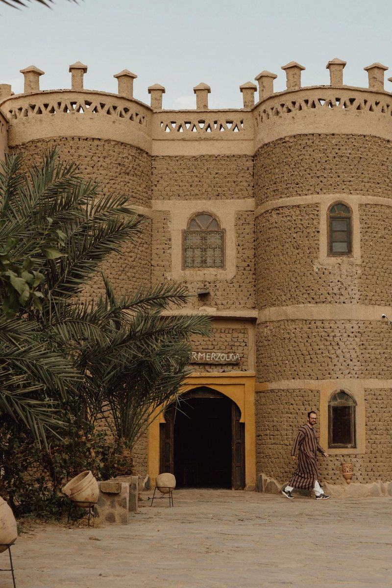 Traditionelles Lehmbau-Hotel mit Rundtürmen in Merzouga, Marokko, vor Palmen und mit einem Mann in Djellaba-Gewand